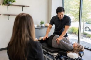 Male chiropractor performing a spinal adjustment on a patient while a female colleague observes in a bright, modern clinic.