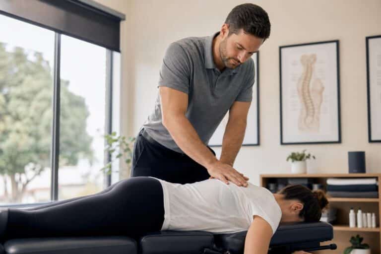 Male chiropractor in a grey polo shirt performing a spinal adjustment on a female patient in a bright, modern clinic with anatomical posters.
